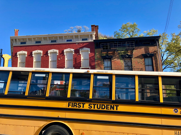 Gelber Schulbus mit der Aufschrift "First Student" vor einem roten Backsteingebäude geparkt, mit Menschen drinnen, Bäumen und einem klaren blauen Himmel im Hintergrund.