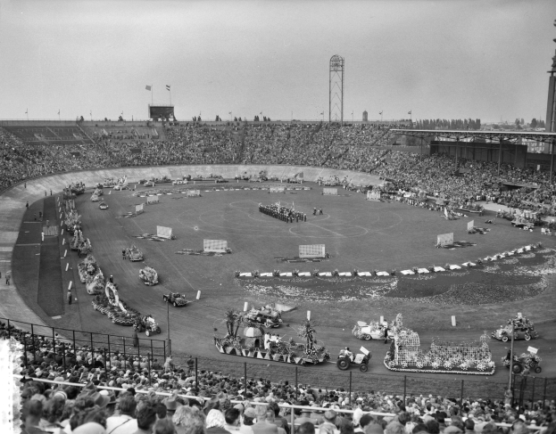 Schwarzes Bild einer großen Menschenmenge, die ein Rennen in einem Stadion mit Fahrzeugen, Zuschauern, Absperrungen, Pfählen, Fahnen, Türmen, Bäumen und Himmel sieht.