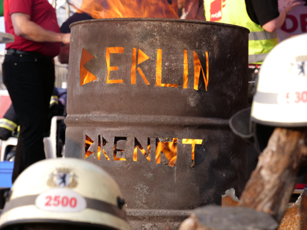 Eine Gruppe von Menschen um ein Feuerhydranten-Schild mit der Aufschrift "Berlin", mit einem brennenden Fass im Vordergrund, Helme unten und einem hölzernen Objekt rechts.