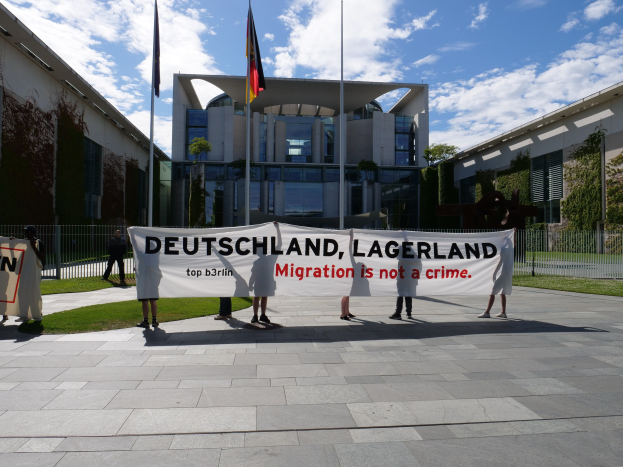 Gruppe von Menschen mit einem Banner, auf dem "Deutschland, Lagerland Migration ist kein Verbrechen" steht, vor einem Gebäude mit Fenstern, umgeben von Gras, einem Metallzaun, Pflanzen, Bäumen und Fahnenmasten unter einem bewölkten Himmel.