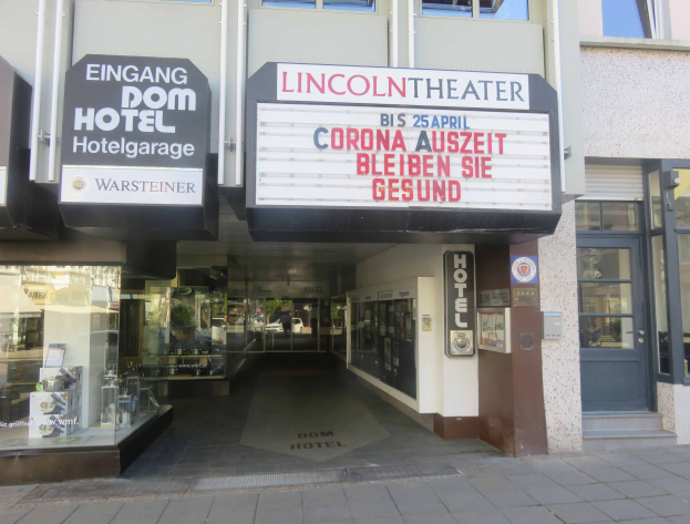 Außenansicht des Lincoln Theaters in Berlin, Deutschland, mit Glasfenstern und -türen und einer Tafel mit Text sowie einem belebten Inneren durch den Eingang sichtbar.
