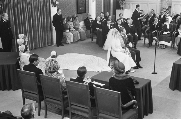 Schwarzes und weißes Foto einer Hochzeitszeremonie mit sitzenden Gästen, einem Mann mit Mikrofon, einer Frau mit einem Bukett, einem Podium mit Mikrofon, Blumenarrangements, einem Vorhang, einem gerahmten Foto an der Wand und einer Tür im Hintergrund.
