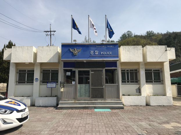 Eine Polizeistation mit einem geparkten Auto davor, das Fenster, eine Tür und eine Tafel mit Text auf dem Gebäude zeigt, mit Fahnen, einem Strommast mit Drähten, Bäumen im Hintergrund und einem bewölkten Himmel.