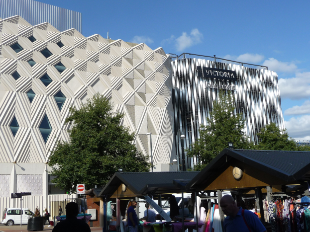 Außenansicht des Victoria and Albert Museums in Birmingham, England, mit Menschen in der Nähe von Kleiderständen, Bäumen, Gebäuden und einem bewölkten Himmel sowie einem Schild im Vordergrund.
