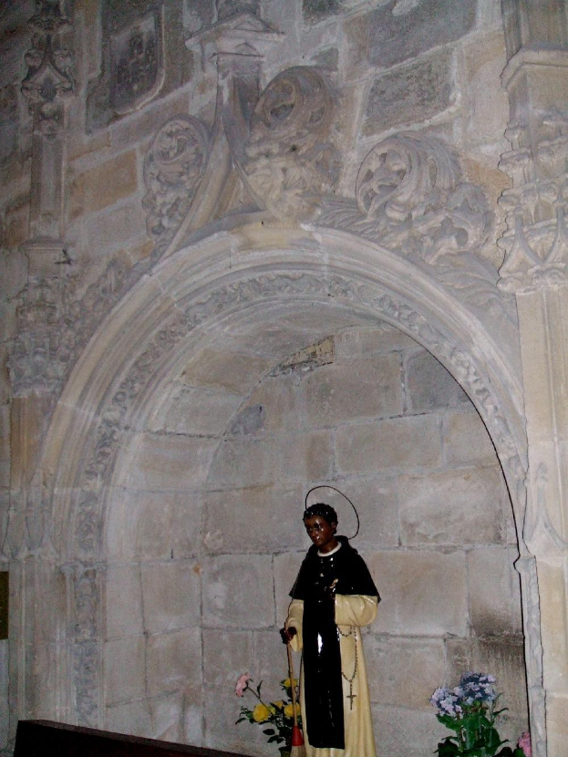 Ein Priester in schwarzer und cremefarbener Robe steht in einem Kirchenbogen, hält einen Stab, mit blühenden Pflanzen daneben und intriganten Schnitzereien an den Wänden dahinter.