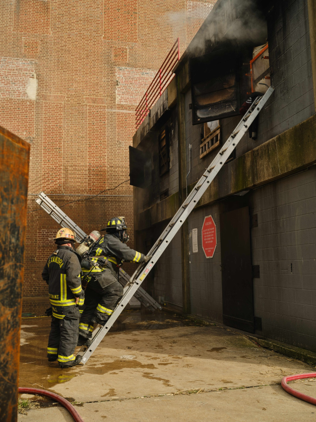 Feuerwehrleute mit Helmen klettern an einer Leiter zu einem raucherfüllten Gebäude hoch, mit einem metallischen Objekt links und Rohren am Boden.