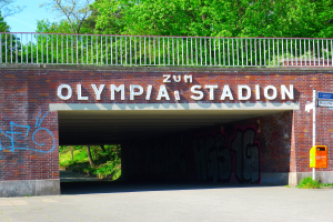 Der Eingang zum Olympiastadion in Berlin, Deutschland, mit einer Brücke, einem Metallzaun, einer Schautafel, einer Box, Pflanzen, Gras, einer Baumgruppe und einem bewölkten Himmel.