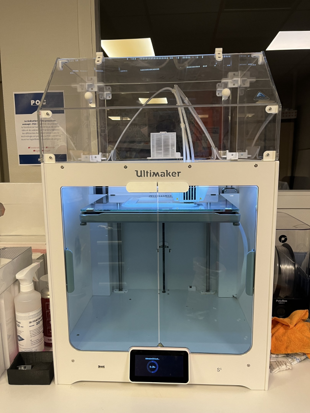 Ein 3D-Drucker, der als Ultimaker etikettiert ist, steht auf einem Schreibtisch neben einem Computer, mit einem Mobiltelefon, einem Tuch und anderen Gegenständen auf dem Tisch, vor einer Wand im Hintergrund.