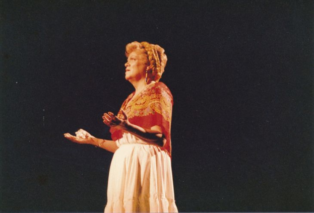 Eine Frau in einem weißen, langärmligen Kleid mit hohem Kragen steht auf einer Bühne mit ausgestreckten Händen, ihr Haar ist zu einem Dutt gebunden, vor einem dunklen Hintergrund.