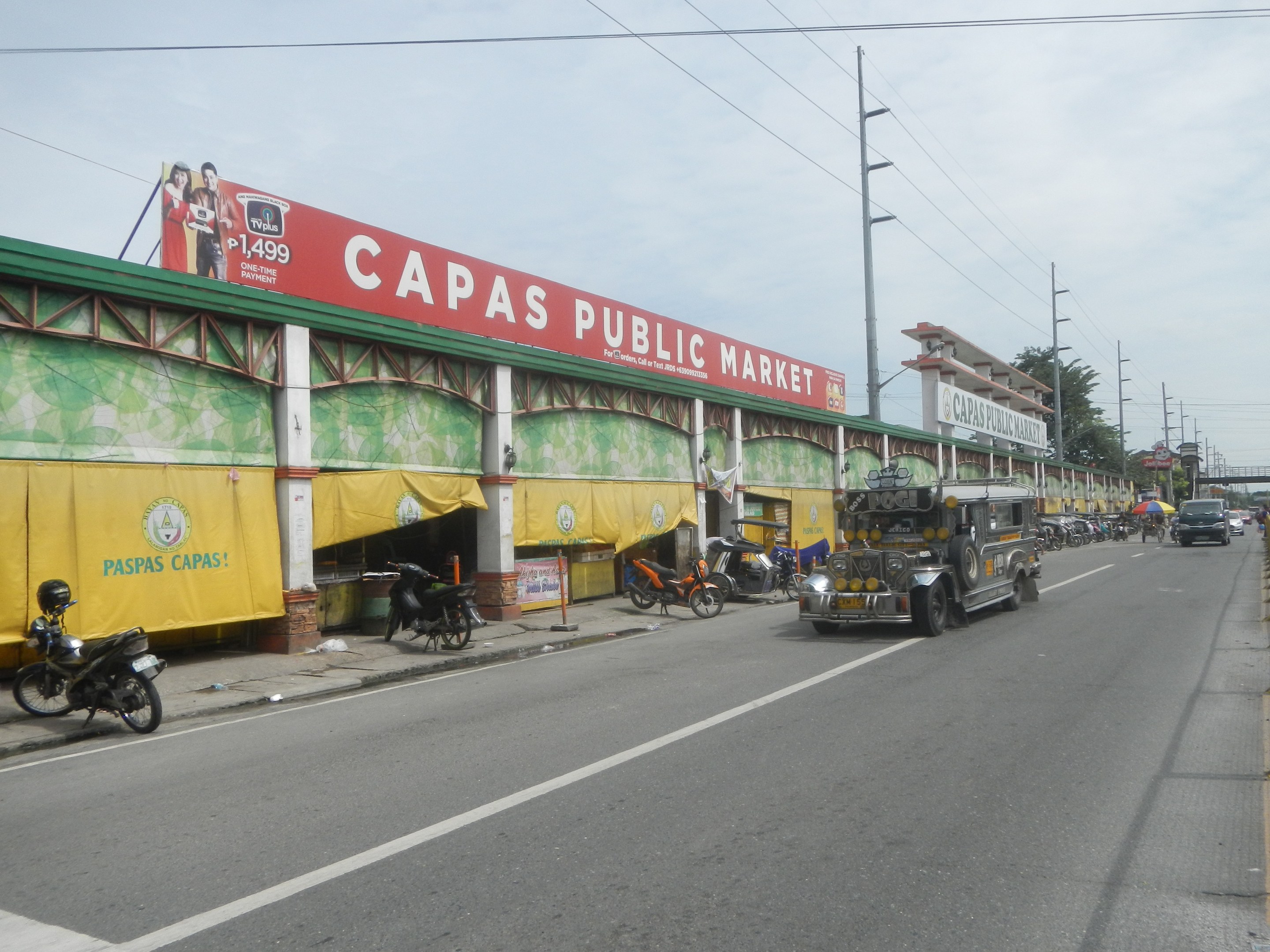 Eine belebte Stadtstraße mit Fahrzeugen, einem Fußweg, Strommasten, Gebäuden, Bäumen und einem bewölkten Himmel, mit einem Gebäude mit der Aufschrift "Capas Public Market" im Vordergrund.