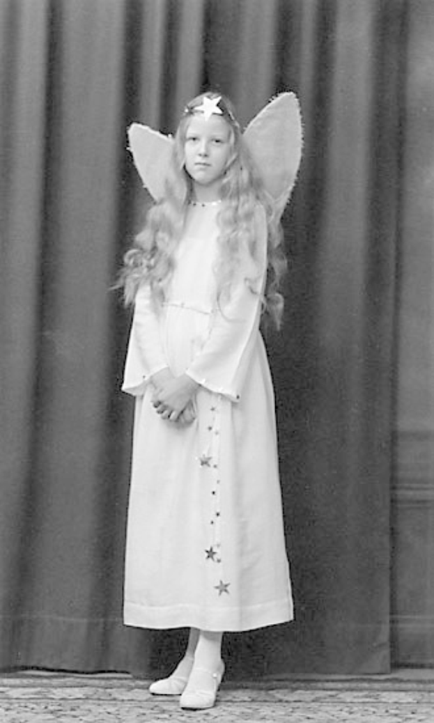 Schwarz-weiß-Foto eines jungen Mädchens in einem weißen Kleid mit Engelsflügeln auf einer Bühne vor einem Vorhang, das Schuhe trägt.