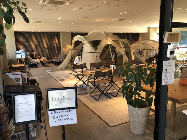 Ein Restaurant von außen in Tokyo mit Tischen, Stühlen und Topfpflanzen in der Nähe einer Glastür, das ein paar sitzende und stehende Menschen sowie Zelte und Deckenleuchten im Hintergrund zeigt.