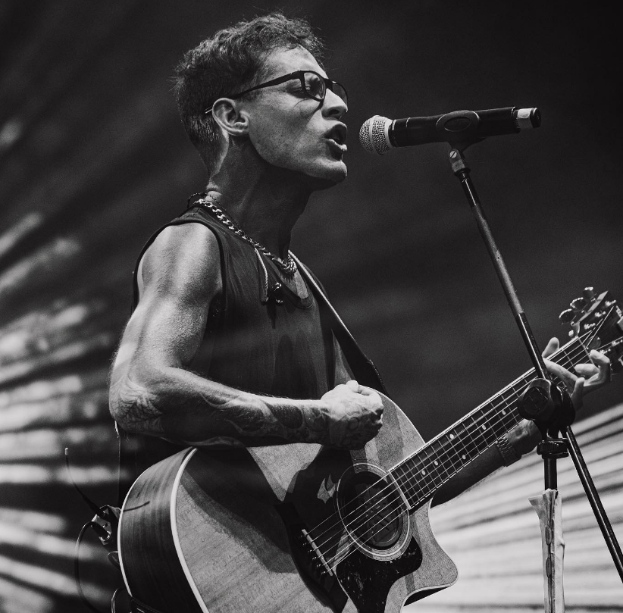 Ein Mann mit Brille spielt Gitarre und singt in ein Mikrofon auf der Bühne der O2 Academy Brixton, dargestellt in Schwarz-Weiß.