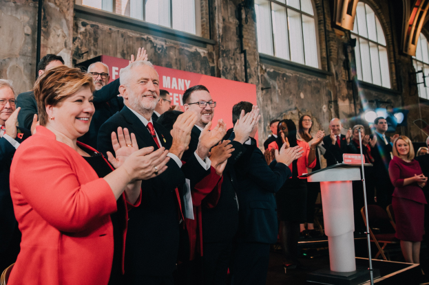 Eine Gruppe von Menschen, die vor einer Menge stehen und applaudieren, mit einem Podium, einem Mikrofon und einer Tafel mit Text auf der rechten Seite und einem Banner, Stühlen, Fenstern und Lichtern im Hintergrund.