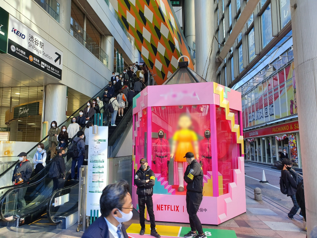 Eine Gruppe von Menschen steht vor einem Gebäude mit Glaswänden und Texttafeln, einige tragen Masken, während eine Rolltreppe mit Menschen daneben fährt; ein pinker Kasten in der Mitte zeigt Schaufensterpuppen und trägt 'Netflix's new Netflix booth in Tokyo'.