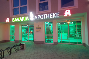 Ein Gebäude mit Glastüren und Fenstern, beleuchtet von Lichtern, hat ein Schild mit der Aufschrift "Bavaria Apotheke"; drinnen sind Regale mit verschiedenen Gegenständen zu sehen, und einige Gegenstände liegen links auf dem Boden.