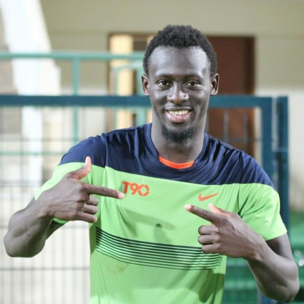 Ein Mann in einem grünen und blauen Hemd, wahrscheinlich Emmanuel Adebayor, zeigt mit einem Lächeln auf die Kamera, mit einem unscharfen Geländer, Wand und Tür im Hintergrund.