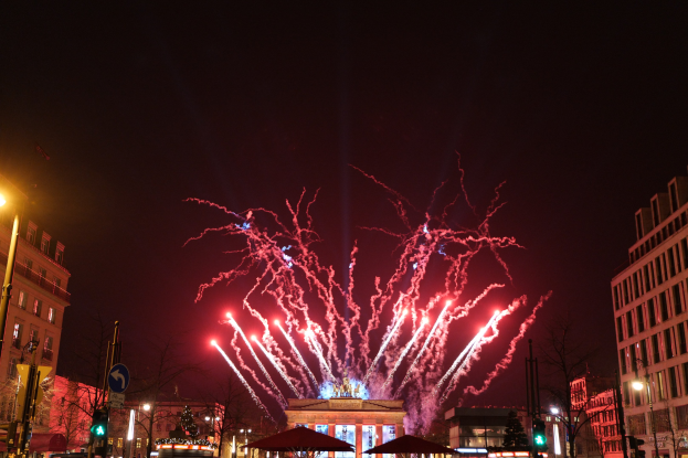 Eine belebte Stadtstraße während eines Silvesterfeuerwerks in Berlin mit Menschen, Fahrzeugen, Bäumen, Straßenmöbeln und beleuchteten Gebäuden unter Feuerwerk am Himmel.