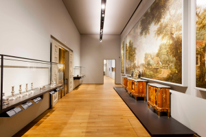 Ein Raum im Museum of Fine Arts in Amsterdam, der verschiedene Kunstwerke, darunter Gemälde, Objekte, Texttafeln und eine Glasbox mit Gegenständen darin, zeigt, beleuchtet von Deckenlampen.