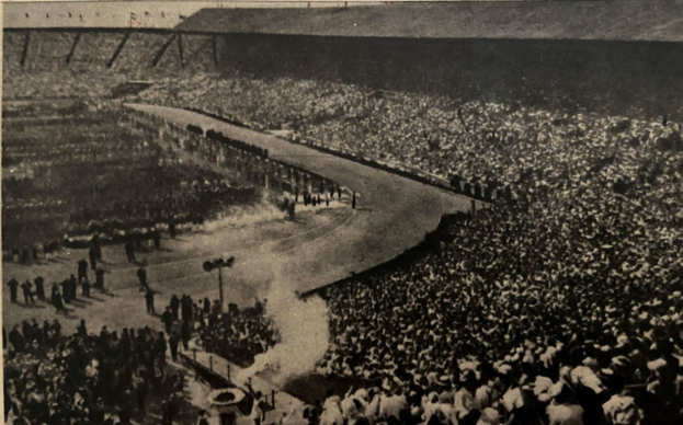 Schwarzes und weißes Foto einer großen Menge in einem Stadion mit einer zentralen Straße und Pfosten auf beiden Seiten, das Text am unteren Rand enthält.