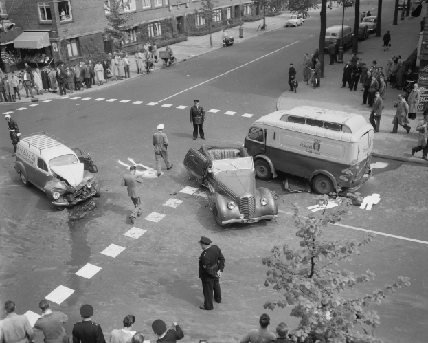 Ein Schwarz-Weiß-Bild einer Autounfall-Szene an einer Straßenkreuzung mit mehreren Fahrzeugen, darunter ein Van, und einer Gruppe von Menschen drumherum, mit Gebäuden, Bäumen, Pfählen und Texttafeln im Hintergrund.