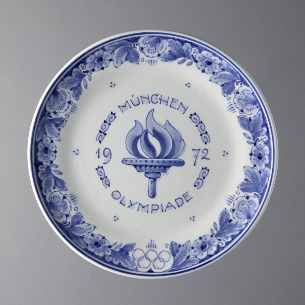 Ein blau-weißer Teller mit dem Olympischen Logo und detaillierten Designs, der die Olympischen Spiele in München feiert, vor einem grauen Hintergrund.