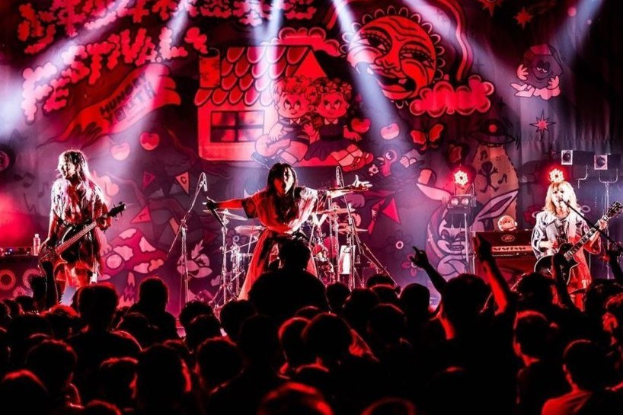 Ein Live-Auftritt der japanischen Rockband Korn auf der Bühne, mit Musikern, die Gitarren spielen und in Mikrofone singen, einem beleuchteten Gemälde an der Wand dahinter und einem sichtbaren Publikum unten.