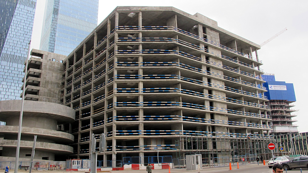 Ein großes Bauwerk im Bau in einer städtischen Umgebung, umgeben von Wolkenkratzern, Fahrzeugen, Fußgängern, Verkehrsinfrastruktur und Baumaschinen unter einem klaren Himmel.