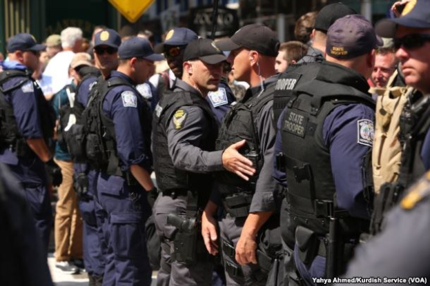 Gruppe von Polizisten in Uniform auf einer Straße stehend, einige mit Waffen, mit Gebäuden und Schildern im Hintergrund.