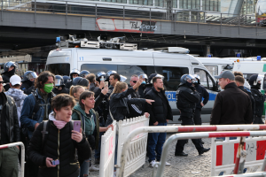 Eine Gruppe von Menschen steht hinter Barrieren und schaut auf Polizeifahrzeuge, einige tragen Helme und halten Telefone, mit einer Brücke und Gebäuden im Hintergrund während einer Demonstration in Berlin.