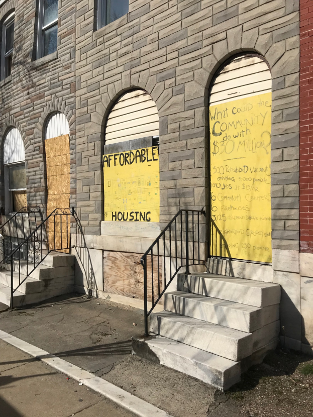 Ein zweistöckiges Gebäude mit vernagelten Fenstern und einem Schild mit der Aufschrift "Bezahlbarer Wohnraum", das eine Treppe mit Geländern zum Eingang führt und eine Straße am unteren Bildrand zeigt.