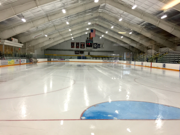 Großes Hallen-Eisstadion mit Deckenbeleuchtung, Werbetafeln, einer Rückwand, Treppen mit Geländern und einigen Menschen, was auf eine Sportanlage hinweist, die likely für Hockey verwendet wird.