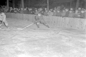 Zwei Männer mit Helmen und Hockeystöcken spielen auf einem Eisstadion, das von einem Holzzaun umgeben ist, vor einem dunklen Hintergrund mit fernen Bäumen in Schwarz-Weiß.