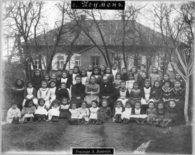 Schwarzes und weißes Foto von Kindern, die vor einem Gebäude mit Fenstern posieren, umgeben von Bäumen und einem klaren Himmel, mit Text unten: "Jugoslawien, die erste Schule der Welt".