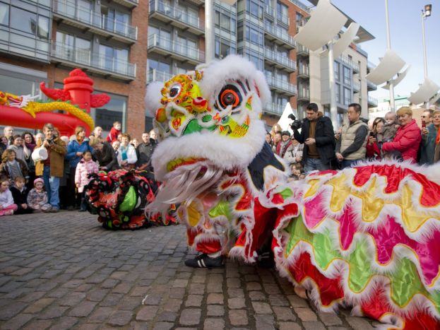 Ein lebendiges chinesisches Neujahrsfest in Amsterdam mit einem Löwen tanzen im Vordergrund, einer Menge Menschen und Gebäuden im Hintergrund unter einem klaren blauen Himmel.