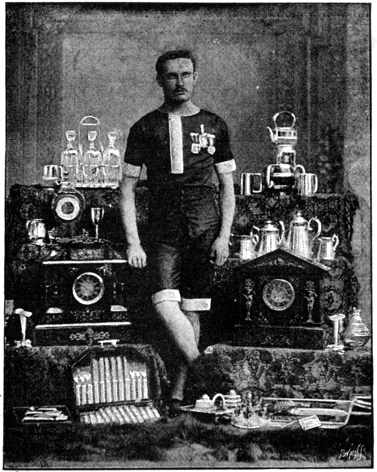 Ein Schwarz-Weiß-Foto eines Mannes in einem Fußballtrikot, der vor einem Tisch mit Pokale und Trophäen steht, mit der Aufschrift "1909-1910-Weltmeisterschaftssieger" unten.