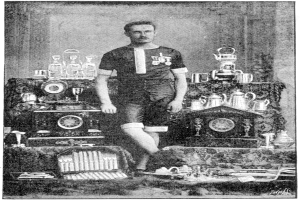 Ein Schwarz-Weiß-Foto eines Mannes in einem Fußballtrikot, der vor einem Tisch mit Pokale und Trophäen steht, mit der Aufschrift "1909-1910-Weltmeisterschaftssieger" unten.