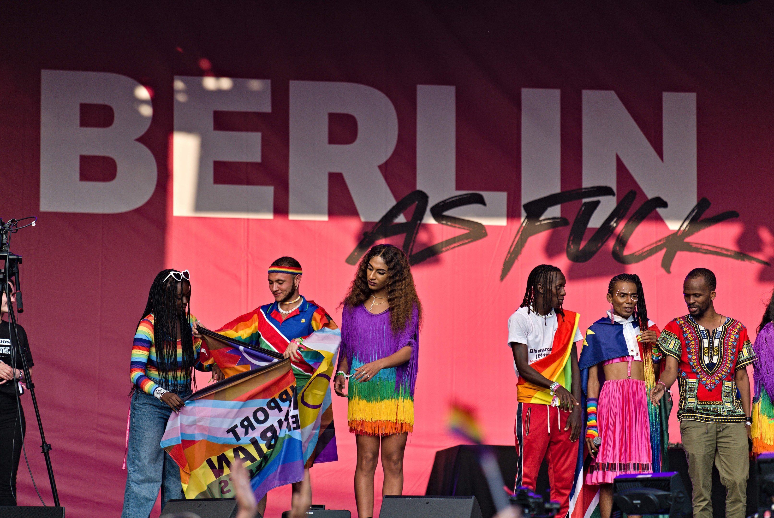 Eine Gruppe von Menschen auf einer Bühne mit einer Frau, die eine Flagge in der Mitte hält, einer Person, die eine Kamera auf einem Stativ links bedient, und einem Banner im Hintergrund mit der Aufschrift "Berlin as Fuck 2019".