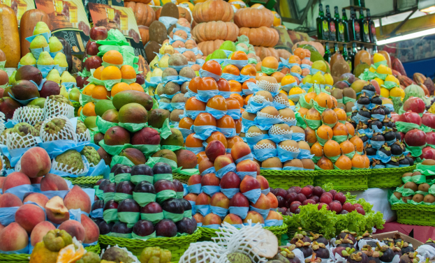 Ein Marktstand mit Äpfeln, Orangen, Birnen und anderem Obst sowie Flaschen auf Regalen unter heller Beleuchtung.