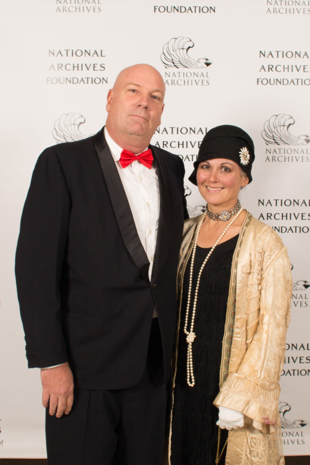 Ein Mann in einem schwarzen Anzug mit einem weißen Hemd und schwarzer Fliege steht neben einer Frau in einem eleganten Kleid mit einem gemusterten Schal auf einem roten Teppich, mit einem weißen Banner mit Text im Hintergrund.