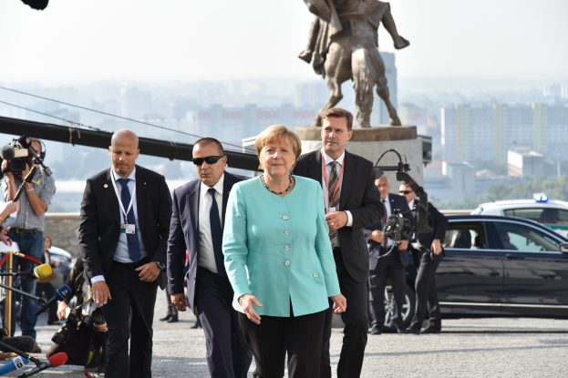 Bundeskanzlerin Angela Merkel und Bundespräsident Joachim Gauck gehen auf einer Straße in Berlin, umgeben von Menschen mit Kameras und Mikrofonen, mit Fahrzeugen, einer Statue, Gebäuden, Bäumen und einem klaren blauen Himmel im Hintergrund.