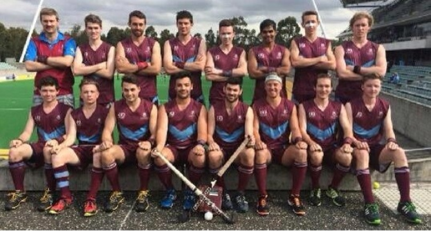 Eine Gruppe von Männern in Sportuniformen steht zusammen auf einem Feld und hält Hockeystöcke, feiert ihren Sieg mit einem Stadion und Zuschauern im Hintergrund unter einem klaren blauen Himmel.