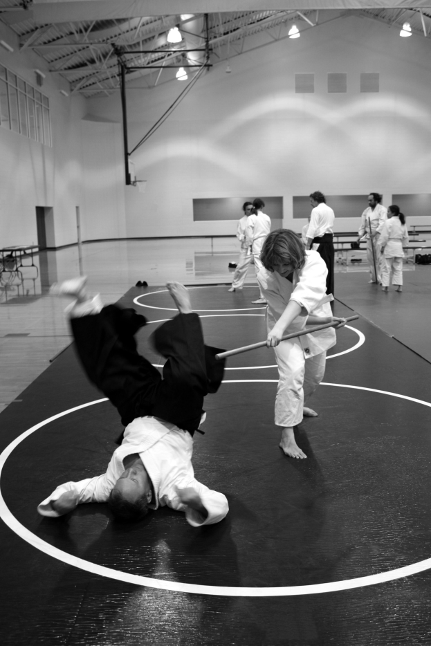 Zwei Personen üben Karate mit Stöcken in einer Turnhalle, während andere zuschauen, mit sichtbaren Deckenleuchten im Hintergrund.