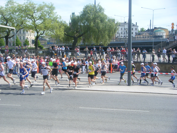 Gruppe von Läufern bei einem Marathon auf einer Straße mit Metallzaun, Zielpfosten, Band, Zuschauern, Barrieren, Pfosten, Schildern, einer Brücke, Gebäuden, Bäumen und einem bewölkten Himmel.