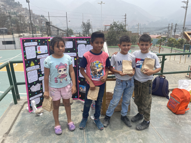 Gruppe von Kindern auf einem Basketballfeld mit Papiertüten in den H├Ąnden, mit Tüten und Gegenst├Ąnden rechts und einer Tafel mit Text links, vor dem Hintergrund von Z├Ąunen, Masten, Stra├čenlaternen, ├Ąltere Strommasten mit Dr├Ąhten, B├Ąumen, Geb├Ąuden, H├Ąngen und Himmel.