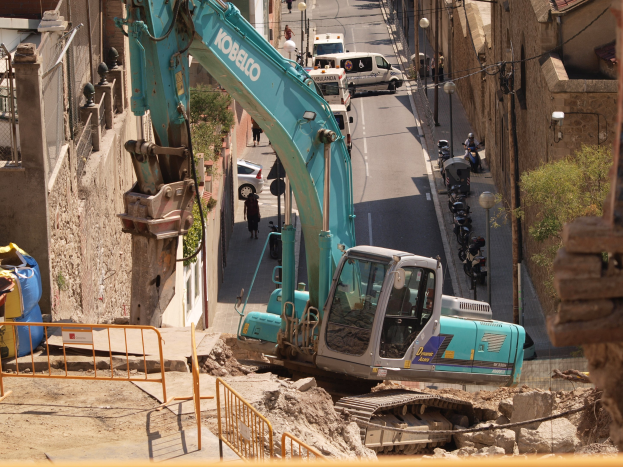 Bagger im Einsatz auf einer städtischen Baustelle mit Gebäuden, Straßeninfrastruktur, Fahrzeugen, Fußgängern und Bäumen.