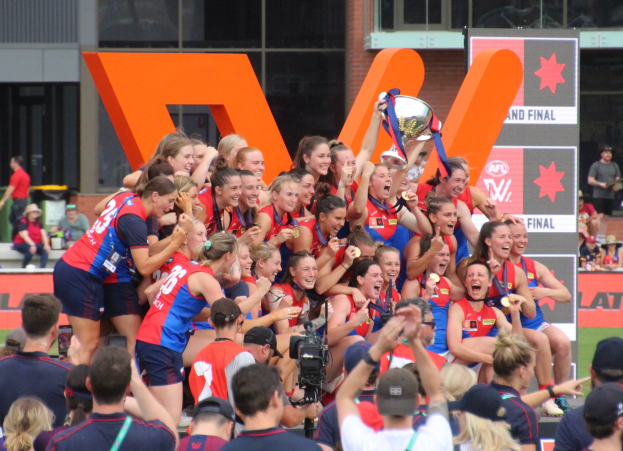 Eine Gruppe von Frauenfußballerinnen in blauen und roten Uniformen feiert mit einem Pokal vor einer jubelnden Menge auf einem Rasenfeld, mit Zuschauern und Werbetafeln im Hintergrund.