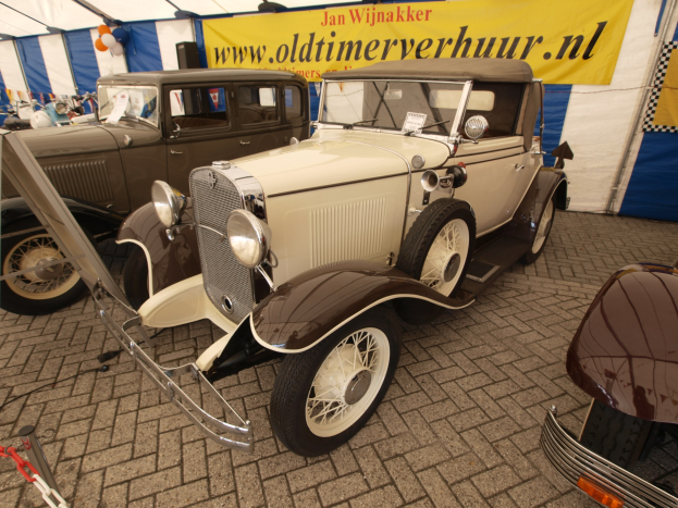 Ein vintage-Rumbelsitz-Auto, das vor einem blauen und weißen Zelt mit einer Fahne und Luftballons im Hintergrund geparkt ist.