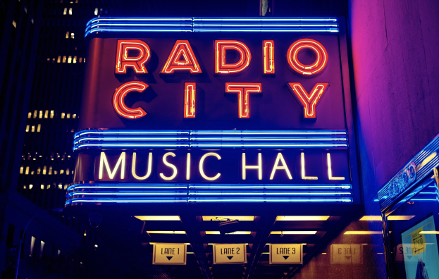 Außensicht des Radio City Music Hall in New York City bei Nacht, hell erleuchtet mit hohen Gebäuden im Hintergrund, mit einem "Radio City"-Schild und mehreren kleineren Schildern an seiner Basis.