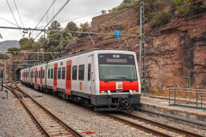 Ein roter und weißer Zug fährt auf Schienen neben einem Berg, mit Strommasten und -leitungen daneben, einer Plattform mit Geländern auf der rechten Seite und Bäumen, Hügeln und einem klaren blauen Himmel im Hintergrund.